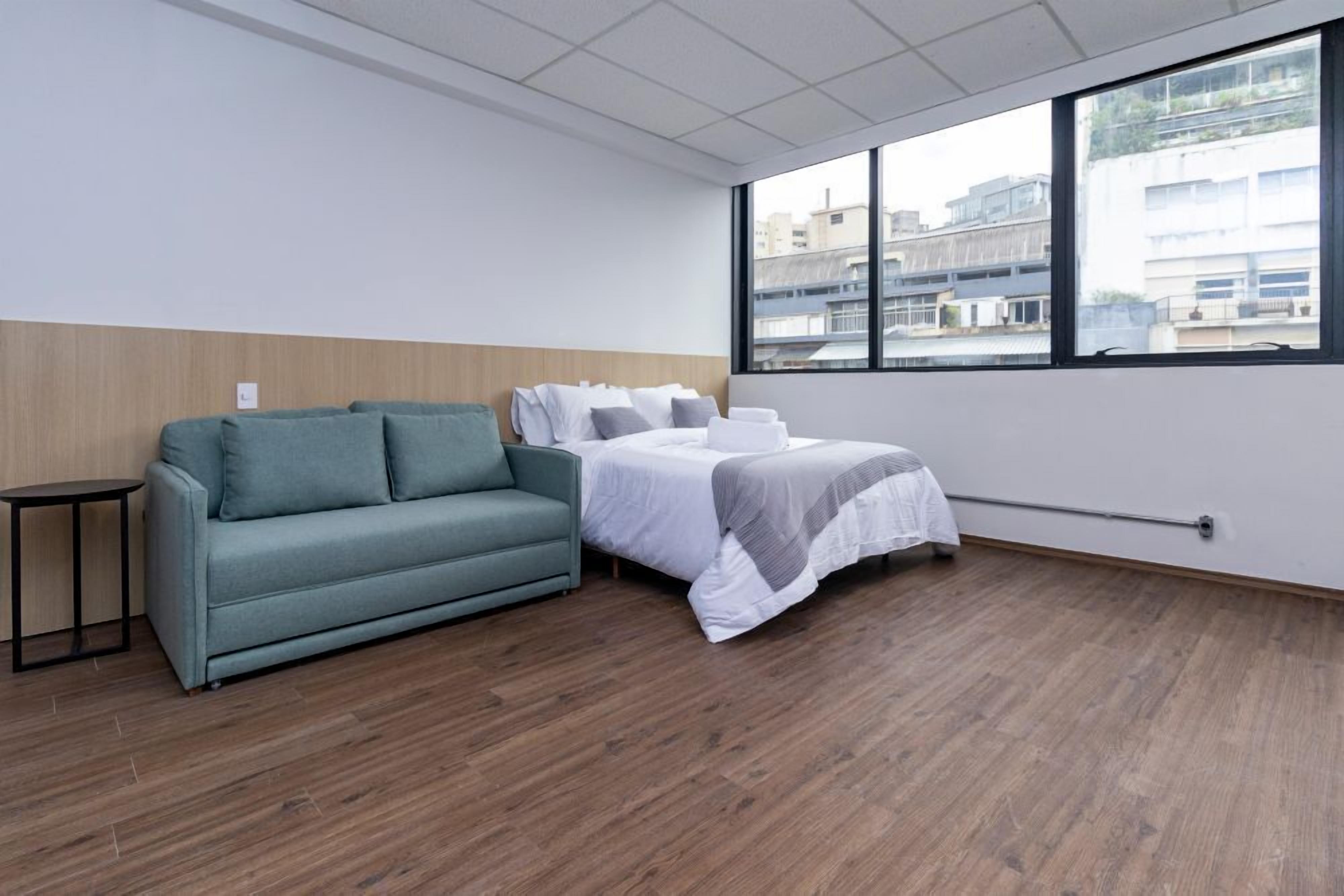 Une chambre d’hôtel avec un lit, un canapé, une petite table et une grande fenêtre donnant sur des immeubles.
