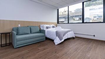 Une chambre d’hôtel avec un lit, un canapé, une petite table et une grande fenêtre donnant sur des immeubles.