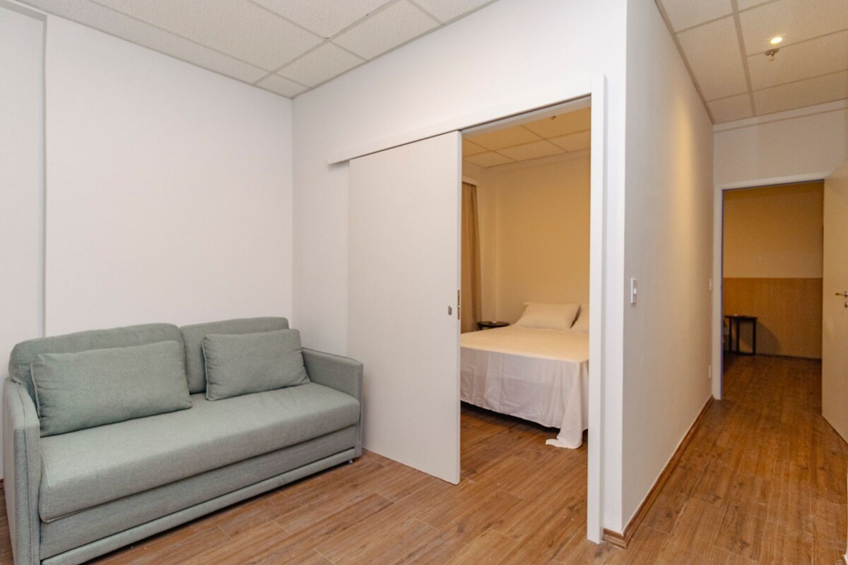 Ein Zimmer mit grauem Sofa, einem Bett mit weißer Bettwäsche und Holzboden.