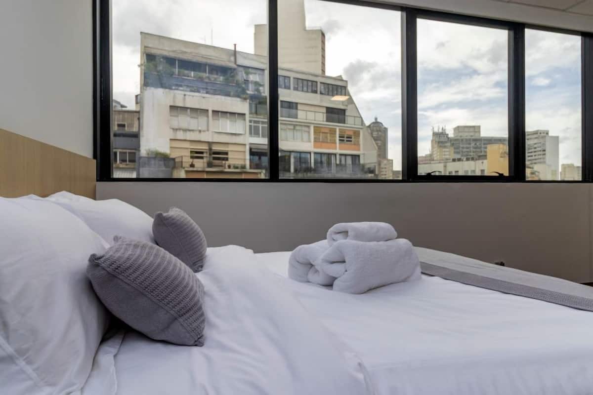 Quarto de hotel com cama, travesseiros, toalha dobrada e uma grande janela com vista para a cidade.