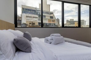 Quarto de hotel com cama, travesseiros, toalha dobrada e uma grande janela com vista para a cidade.