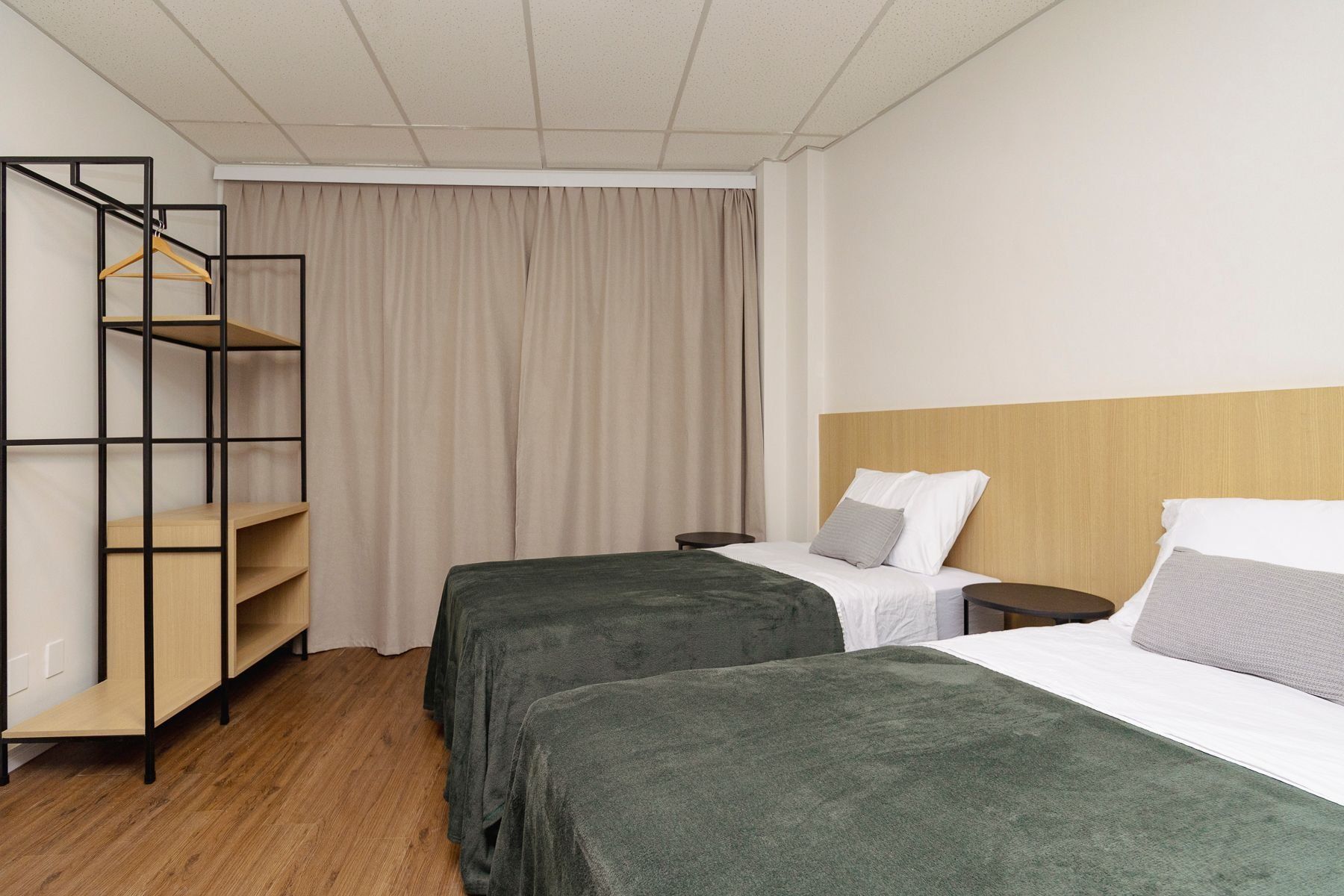 Une chambre d’hôtel avec deux lits, une tête de lit en bois, une armoire avec une tringle pour suspendre les vêtements et un rideau.