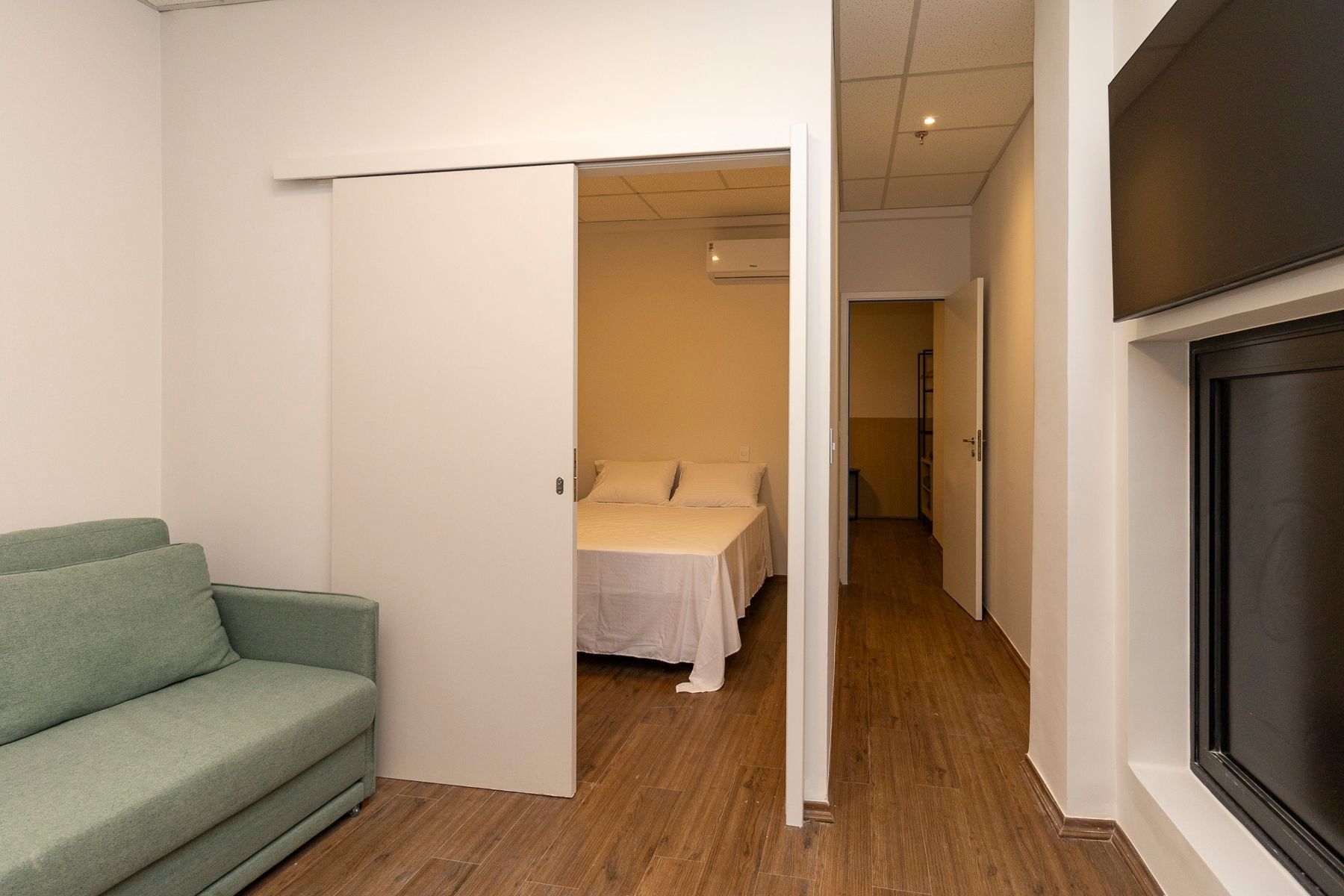 A room with a green sofa, a bed with white bedding, and a television on the wall.