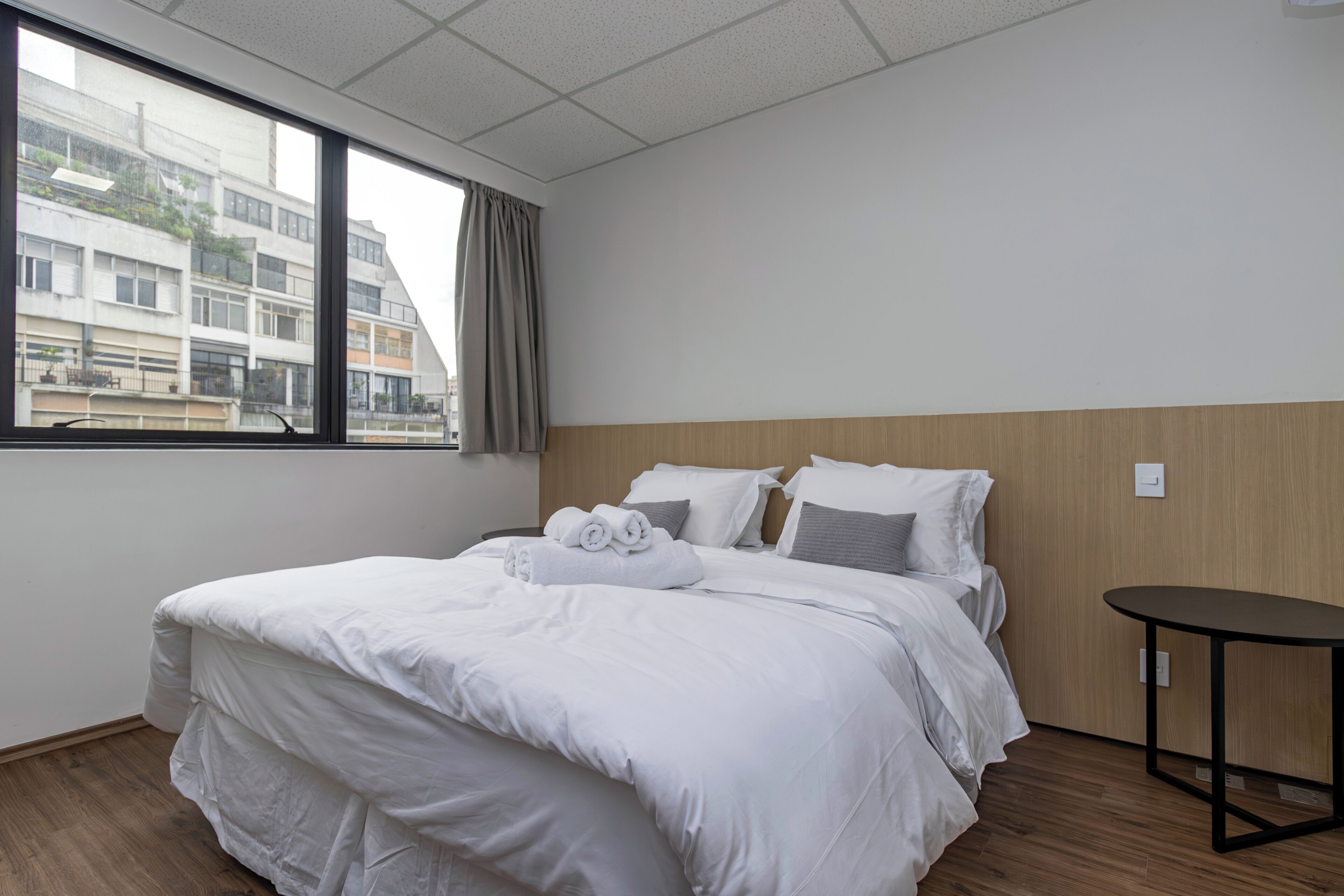A hotel room with a large bed, a small table, a window with a view of buildings, and a wooden headboard.