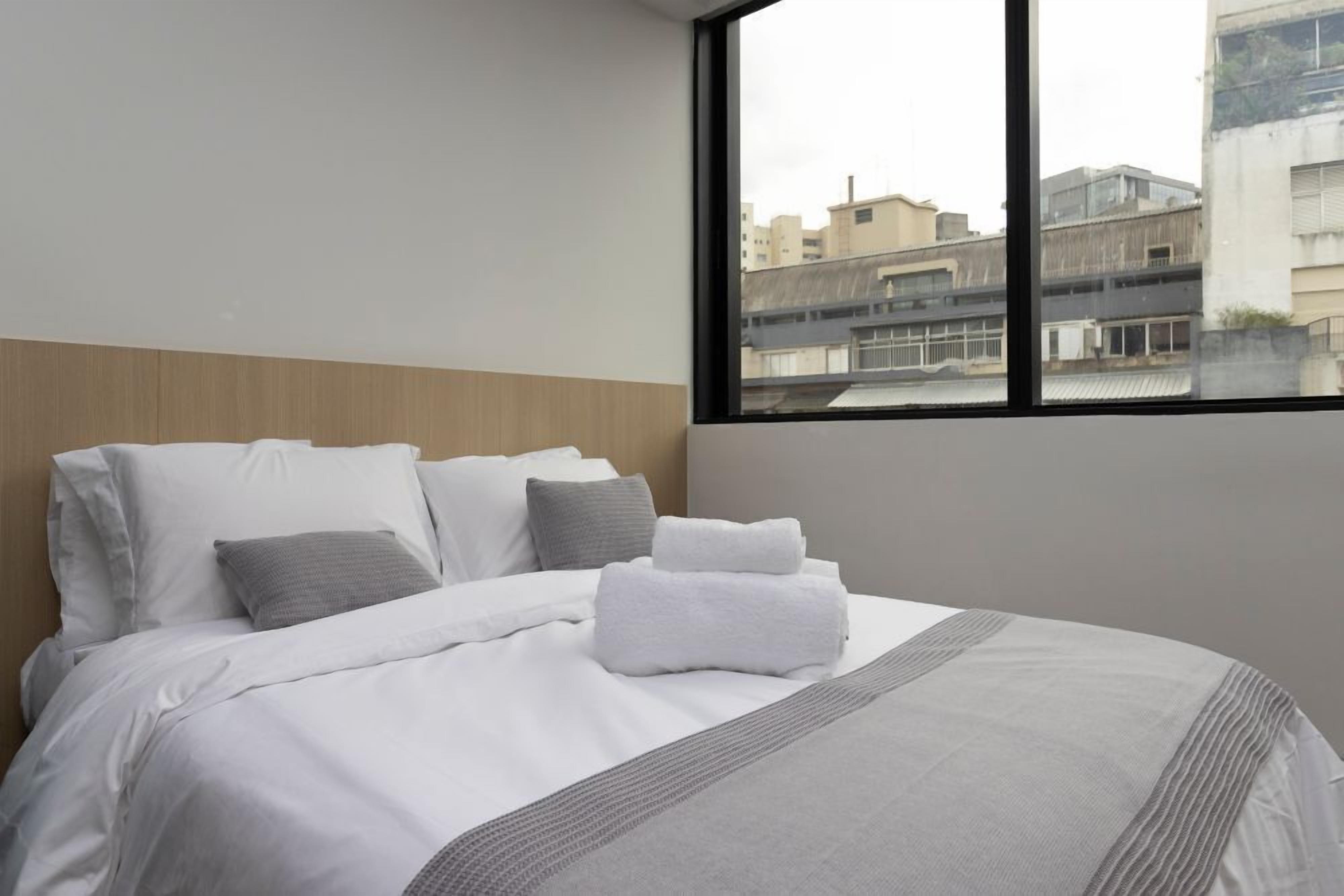 Habitación de hotel con una cama grande, ropa de cama blanca, una manta gris y vistas a edificios a través de una ventana.