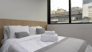 Habitación de hotel con una cama grande, ropa de cama blanca, una manta gris y vistas a edificios a través de una ventana.