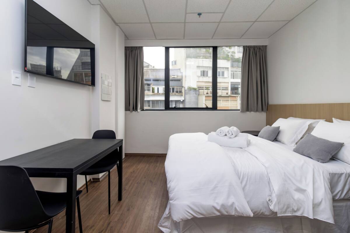 Quarto de hotel com cama, escrivaninha, cadeira, televisão na parede e janela com cortinas.