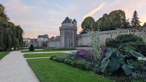 Un château doté d’une tour, entouré d’un jardin bien entretenu où poussent différentes espèces de plantes, et agrémenté d’un chemin pavé.