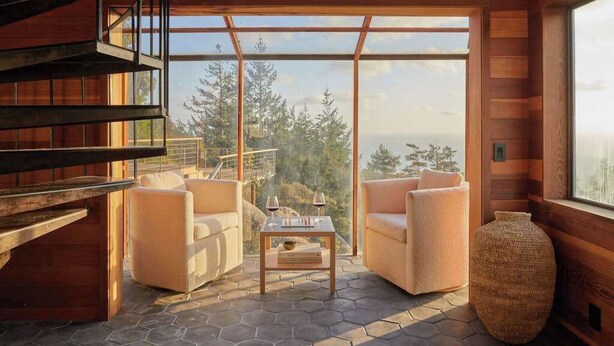 Interior em madeira com sofá, mesa com copos e uma janela ampla com vista para uma floresta.