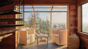A wooden interior with a sofa set, a table with glasses, and a large window overlooking a forest.