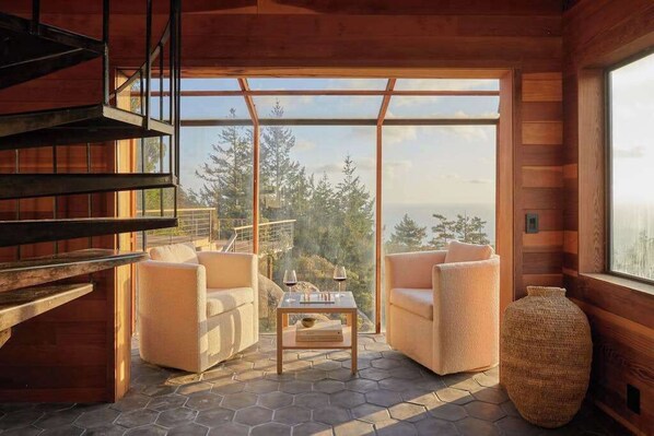 A wooden interior with a sofa set, a table with glasses, and a large window overlooking a forest. - Ocean-view main house and private cottage (Gualala)