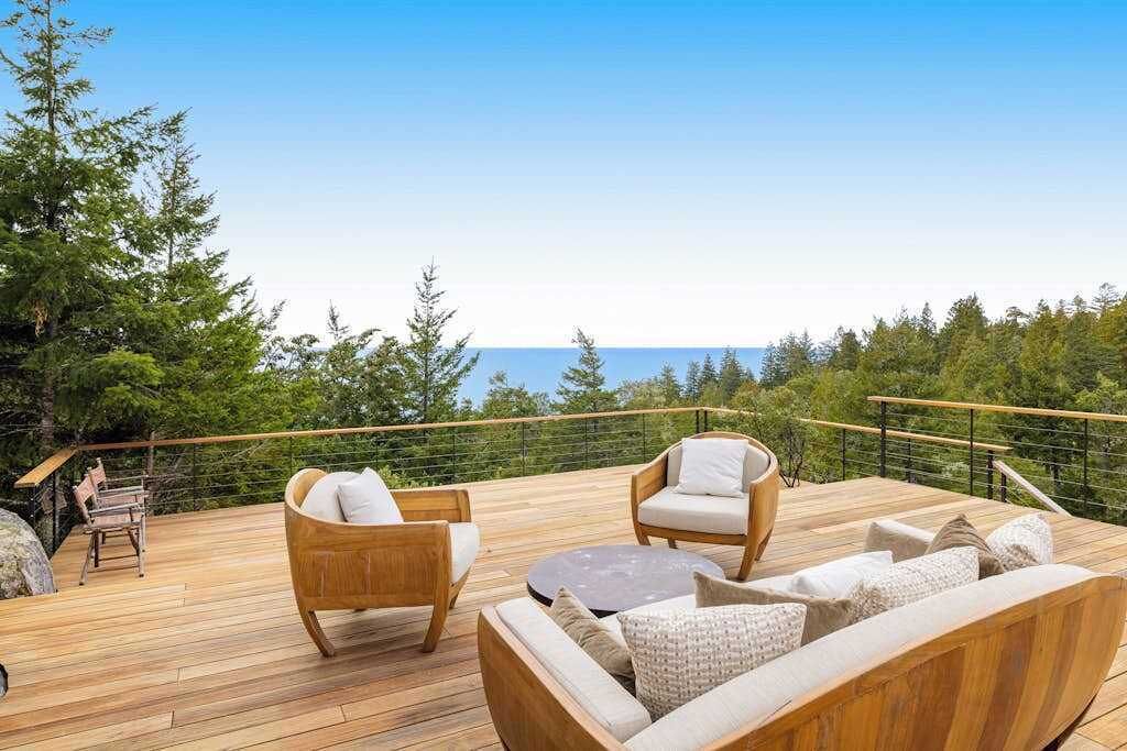 A wooden deck with outdoor furniture, a railing, and a view of the ocean and trees.