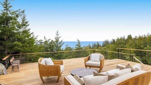 Um deck de madeira com móveis de exterior, guarda-corpo e vista para o oceano e árvores.