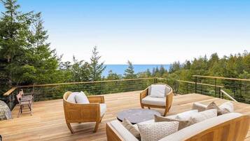 A wooden deck with outdoor furniture, a railing, and a view of the ocean and trees.