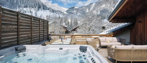 Un jacuzzi con asientos y una mesa, rodeado por una terraza de madera y montañas cubiertas de nieve.