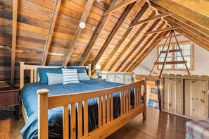 A loft bedroom with a wooden ceiling, a bed with blue bedding, a wooden ladder, and a window.