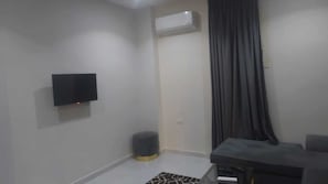 Una habitación con una televisión de pantalla plana montada en la pared, un cubo de basura redondo pequeño, un sofá gris y una alfombra estampada en el suelo.