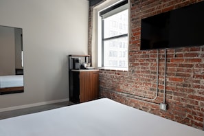 Una habitación con pared de ladrillo, una cama, un televisor montado y una ventana con vistas a edificios.