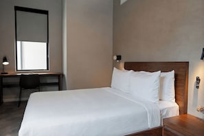 Una cama bien hecha con sábanas blancas, cabecera de madera, un escritorio con silla y una lámpara de pared.