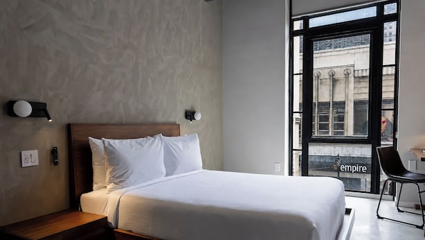 A modern hotel room with a large bed, a nightstand, a wall-mounted light, and a view of the city through a large window. - Sosuite | City View Queen Room w Desk (Philadelphia)