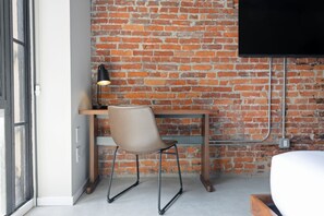 A room with a brick wall, a wooden desk, a chair, a bed, and a TV mounted on the wall.