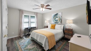 A bedroom with a bed, a desk, a chair, a ceiling fan, a lamp, and a painting on the wall. - Relaxing 2BR 1BA Townhome • Loaded Amenities • Central Location Near I-40 (Jackson)