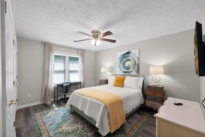 A bedroom with a bed, a desk, a chair, a ceiling fan, a lamp, and a painting on the wall.