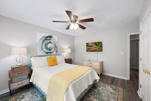 A bedroom with a bed, a ceiling fan, a nightstand with a lamp, a dresser, and a rug.