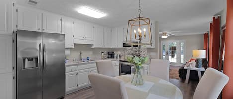 A modern kitchen with white cabinets, a stainless steel refrigerator, a dining table with chairs, and a hanging light fixture.
