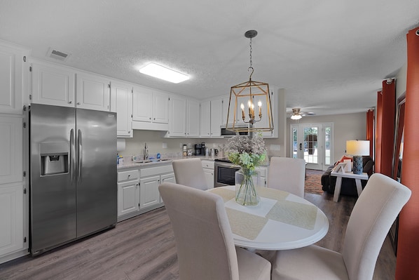 A modern kitchen with white cabinets, a stainless steel refrigerator, a dining table with chairs, and a hanging light fixture.