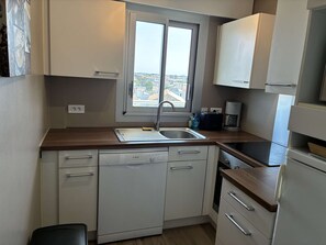 Eine kleine Küche mit Geschirrspüler, Spüle, einem Fenster mit Stadtblick und einer Kaffeemaschine.