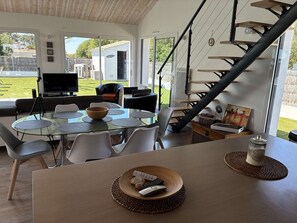 A modern living room with a glass dining table, comfortable seating, a staircase, and a view of the outdoors. - Villa Marthe Océane - Bretignolles sur mer (BRETIGNOLLES SUR MER)