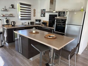 A modern kitchen with a central island, stainless steel appliances, and a dining area with a chair. - Villa Marthe Océane - Bretignolles sur mer (BRETIGNOLLES SUR MER)