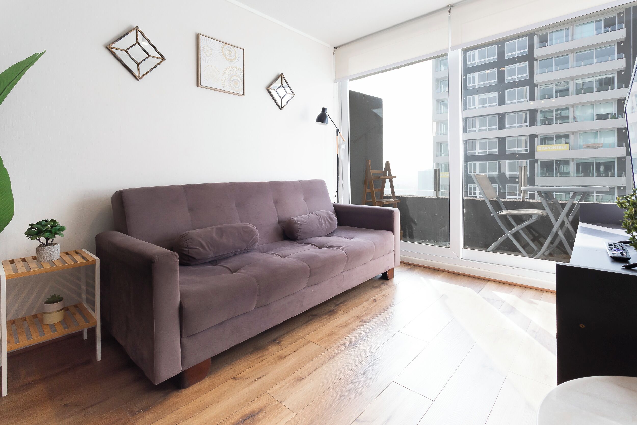 A modern living room with a sofa, a small side table, a potted plant, and a large window with a view of a building.