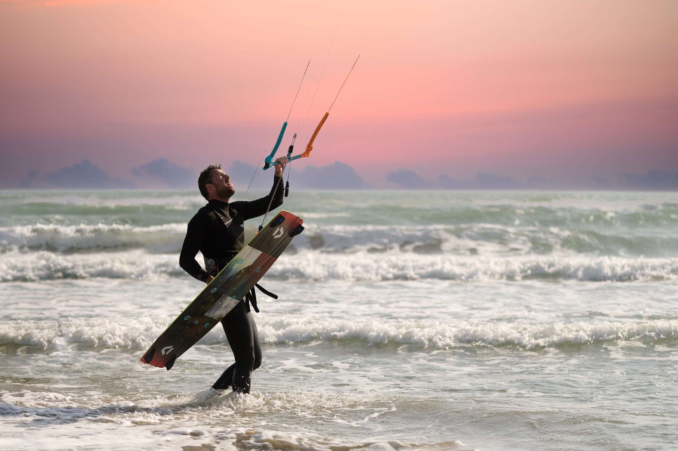 Uma pessoa com fato de neoprene segurando um kite e uma prancha de surf, parada no oceano.