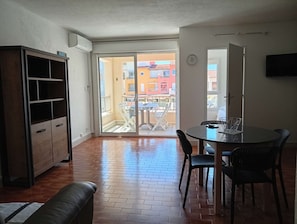 Ein Zimmer mit einer Schiebetür zu einem Außenbereich, einem Esstisch mit Stühlen, einer Couch und einem an der Wand befestigten Fernseher.