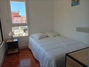 Ein Schlafzimmer mit einem Bett, einem Nachttisch, einer Lampe, einem Fenster und Blick auf Gebäude.