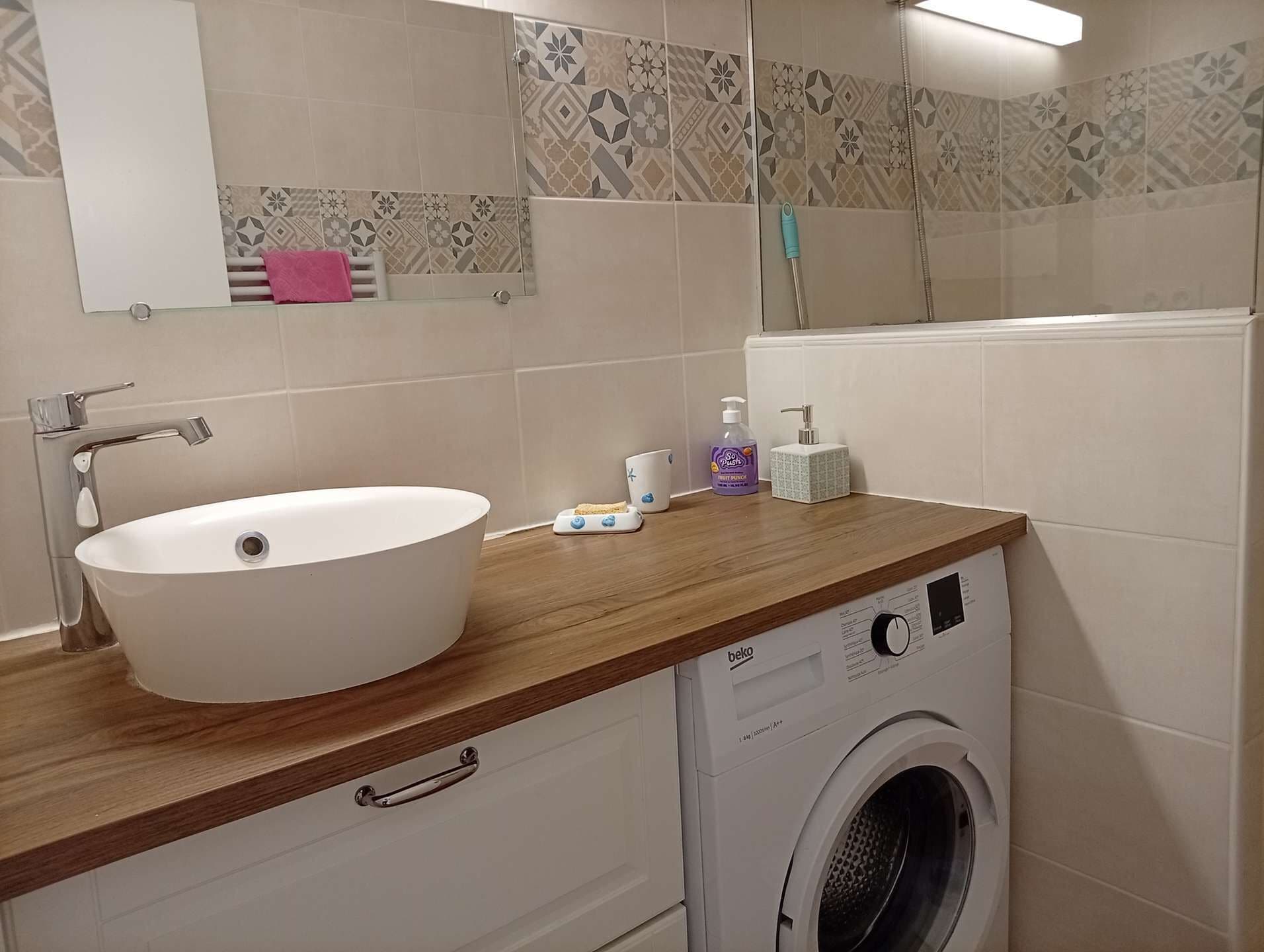Un baño con un lavamanos blanco, encimera de madera, lavadora y azulejos con diseño en las paredes.