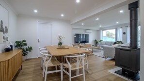 Un vaste espace de vie comprenant une table à manger en bois, des chaises blanches, un canapé, un téléviseur et une cheminée.