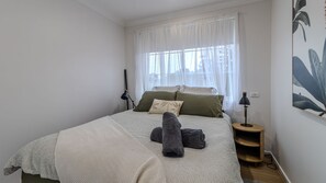 Quarto com cama, almofadas, luminária, mesa de cabeceira e janela com cortinas leves.