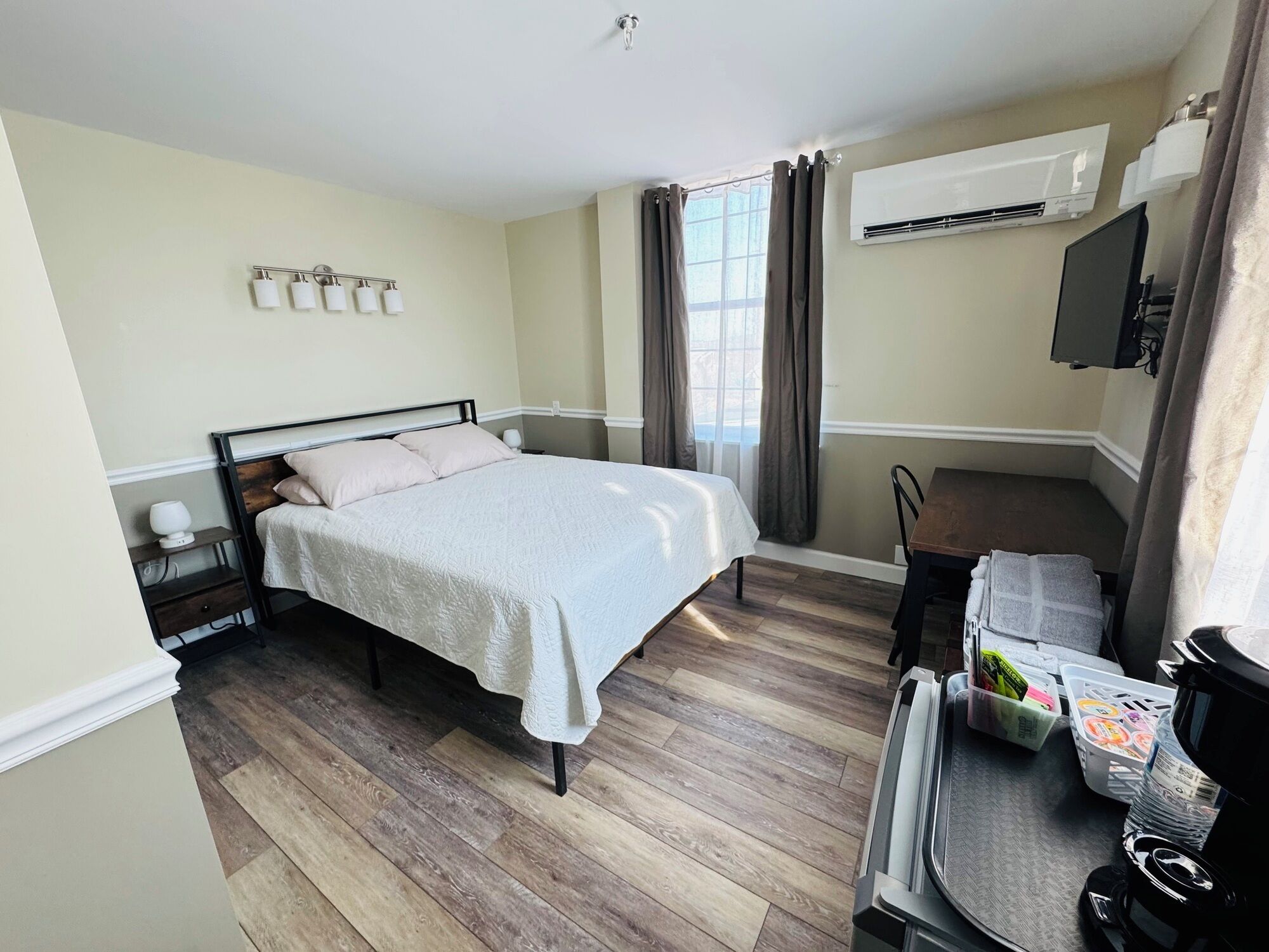A bedroom with a bed, bedside tables, an air conditioning unit, a TV, and a desk with a chair.
