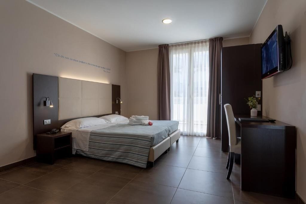 Una habitación de hotel moderna con una cama, un escritorio, una silla, un televisor y un ventanal con cortinas.