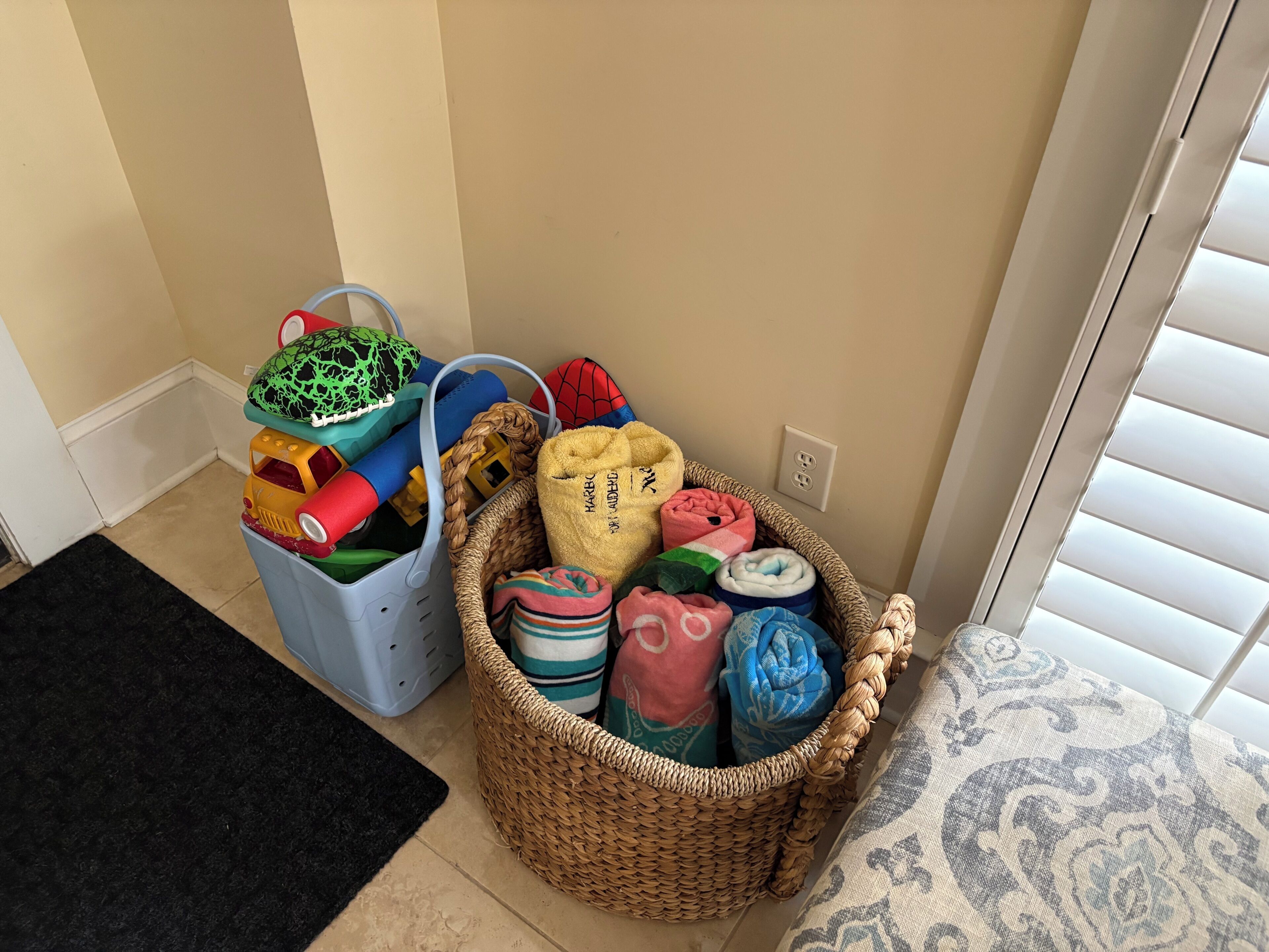 Esquina de una habitación con una cesta de mimbre llena de mantas dobladas y una cesta de plástico con objetos de colores.