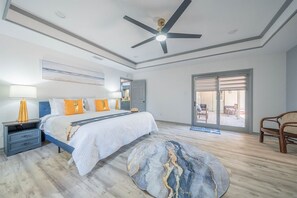 Une chambre à coucher avec un lit, des tables de chevet, un ventilateur de plafond, une grande fenêtre et une vue sur l’extérieur.