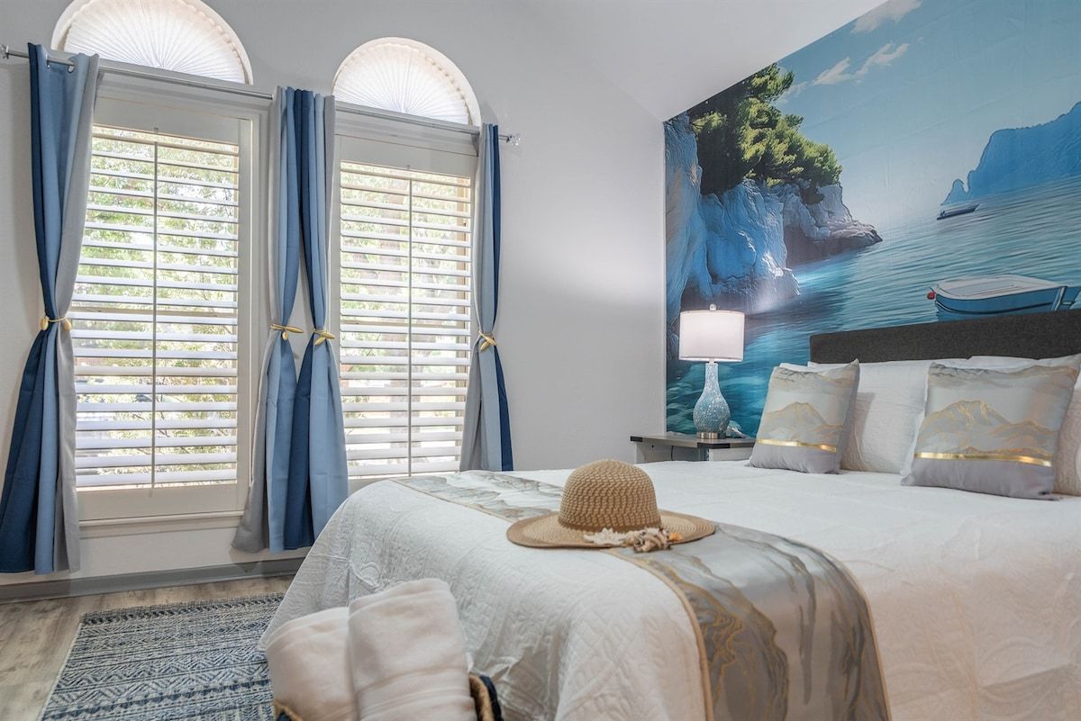 Une chambre à coucher avec un grand lit, un tableau représentant un paysage côtier et une fenêtre avec des rideaux.
