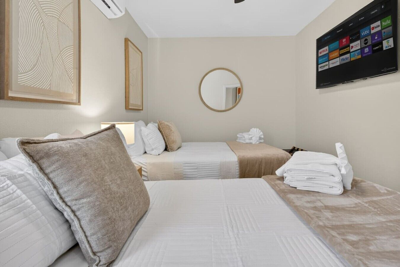Une chambre d’hôtel avec deux lits, un miroir rond, un téléviseur fixé au mur qui permet d’accéder à des services de diffusion en continu, et une œuvre d’art encadrée au-dessus du lit.