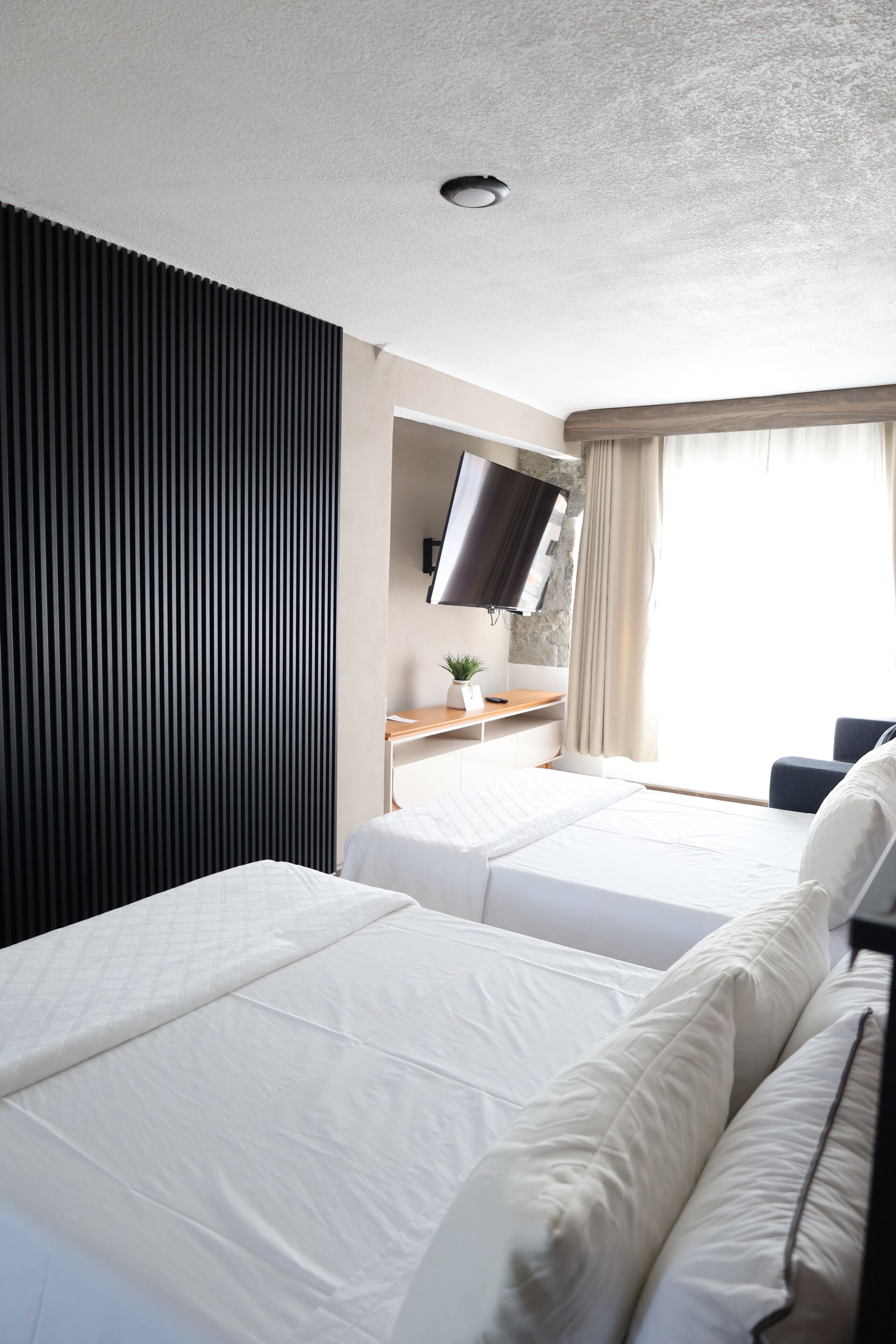 A hotel room with two beds, a flat-screen TV mounted on the wall, a desk with a plant, and a window with curtains.