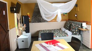 Una habitación con una cama, una mesita con un vaso, una silla, un televisor en la pared y una mosquitera sobre la cama.