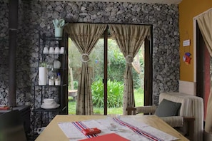 Una habitación con pared de piedra, una cama con colcha a rayas, una mesa de comedor con un objeto rojo y un estante con utensilios de cocina.