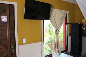 Una habitación con un televisor montado en la pared, una ventana con cortina, una puerta con un letrero y vistas al exterior con vegetación.